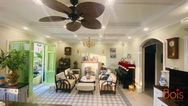 Phòng khách sau thi công: nhóm ghế gỗ trên nền gạch họa tiết, bên cửa xanh và đàn piano.